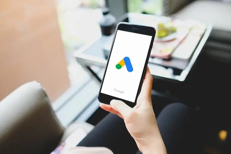 Ao fundo um escritório e no primeiro plano uma mão segurando um celular com a imagem do Google Ads
