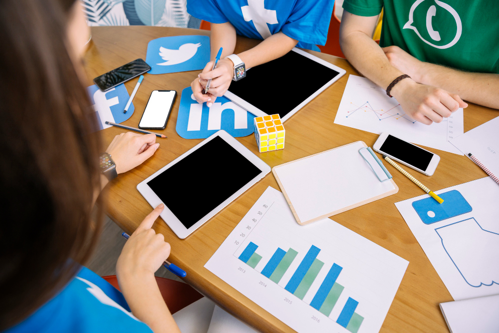 Pessoas reunidas em uma mesa planejando estratégias de marketing digital, com tablets, smartphones, gráficos, anotações e ícones de redes sociais como Twitter, LinkedIn, Facebook e WhatsApp.