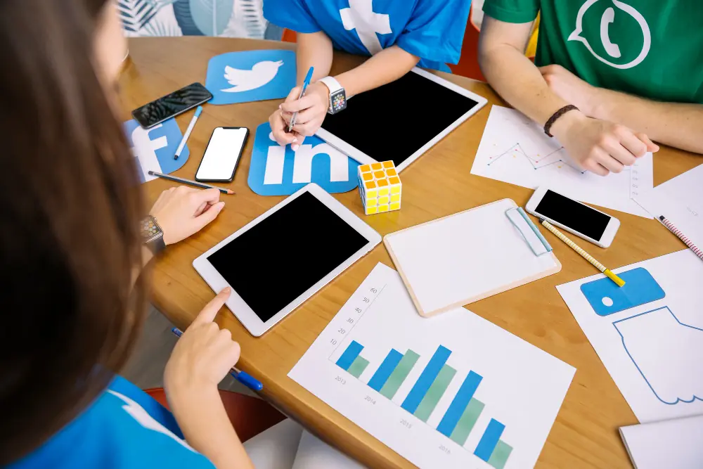 Pessoas reunidas em uma mesa planejando estratégias de marketing digital, com tablets, smartphones, gráficos, anotações e ícones de redes sociais como Twitter, LinkedIn, Facebook e WhatsApp.